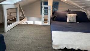 A cozy attic bedroom with a sloped ceiling, a bed with dark bedding, a white throw blanket, and a 'Hello Sun Shine' pillow. - HB Hideaway · 2 Mi to Beach · 60+ Day Savings · Ideal for Snowbirds & WFH! (Huntington Beach)