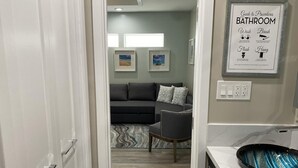 A modern bathroom with a sink, a framed guide to bathroom procedures, and a view into a living area with a sofa and a chair. - HB Hideaway · 2 Mi to Beach · 60+ Day Savings · Ideal for Snowbirds & WFH! (Huntington Beach)