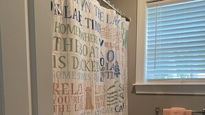 A bathroom with a shower curtain featuring various words and phrases, a towel rack with a towel, and a window with blinds. - Sebago Lake, Maple Cove, Lake House (Casco)
