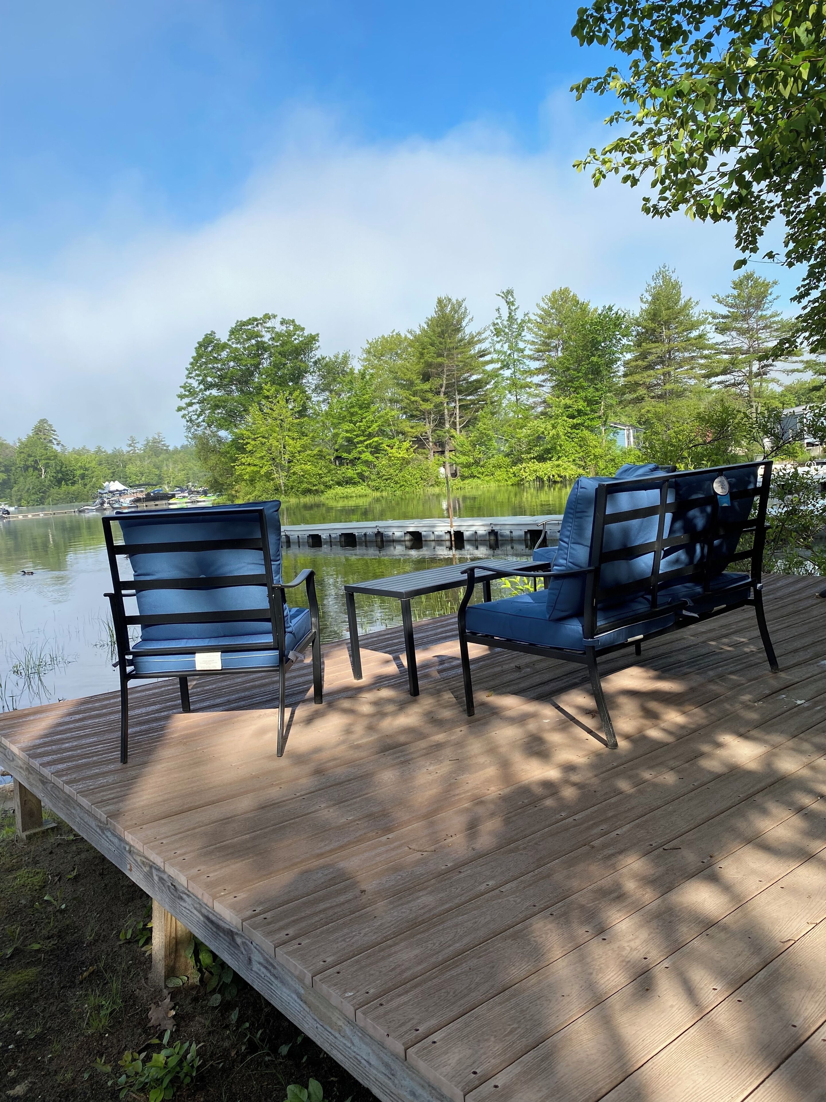 A wooden deck with two blue chairs and a table, overlooking a calm lake with trees in the background.