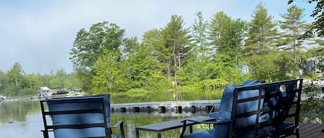 Une terrasse en bois avec deux chaises bleues et une table, donnant sur un lac paisible, avec des arbres en arrière-plan.