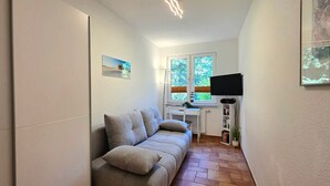 Ein schmaler Flur mit einer Couch, einem Heizkörper, einem Fernseher und einem Fenster mit Jalousien.