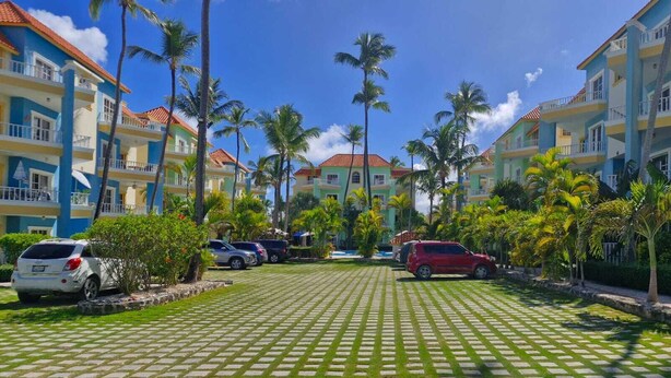 A hotel with a checkered courtyard, palm trees, and parked cars. - Lovely Spacious Condo with Pool View, Close to Beaches and Shops! (Punta Cana)