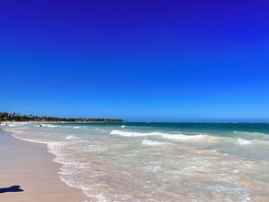 Ein Strand mit klarem, blauem Wasser, einem sandigen Ufer und einer fernen Küstenlinie mit Gebäuden.
