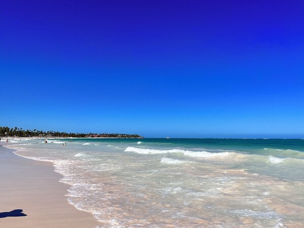 A beach with clear blue water, a sandy shore, and a distant shoreline with buildings.