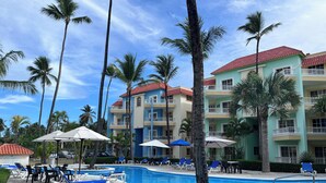 A swimming pool area with lounge chairs and umbrellas, surrounded by palm trees and a multi-story building with balconies.