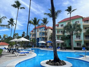 A swimming pool area with lounge chairs and umbrellas, surrounded by palm trees and a multi-story building with balconies.
