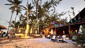 A beachside restaurant with outdoor seating, palm trees, and string lights.
