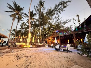 A beachside restaurant with outdoor seating, palm trees, and string lights.
