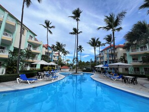 Área de piscina em resort, com várias espreguiçadeiras e guarda-sóis, cercada por palmeiras e edifícios com varandas.