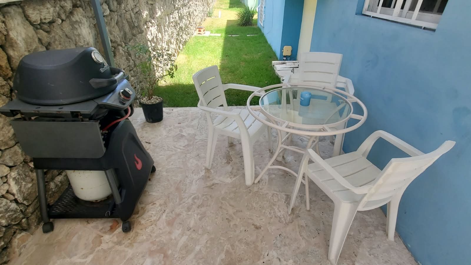 Um pátio com churrasqueira, cadeiras de plástico branco e uma mesa de vidro.