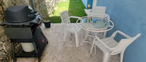 Um pátio com churrasqueira, cadeiras de plástico branco e uma mesa de vidro.