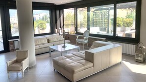 Een moderne woonkamer met een witte bank, een glazen salontafel en een witte fauteuil. Grote ramen bieden uitzicht op het omliggende landschap.