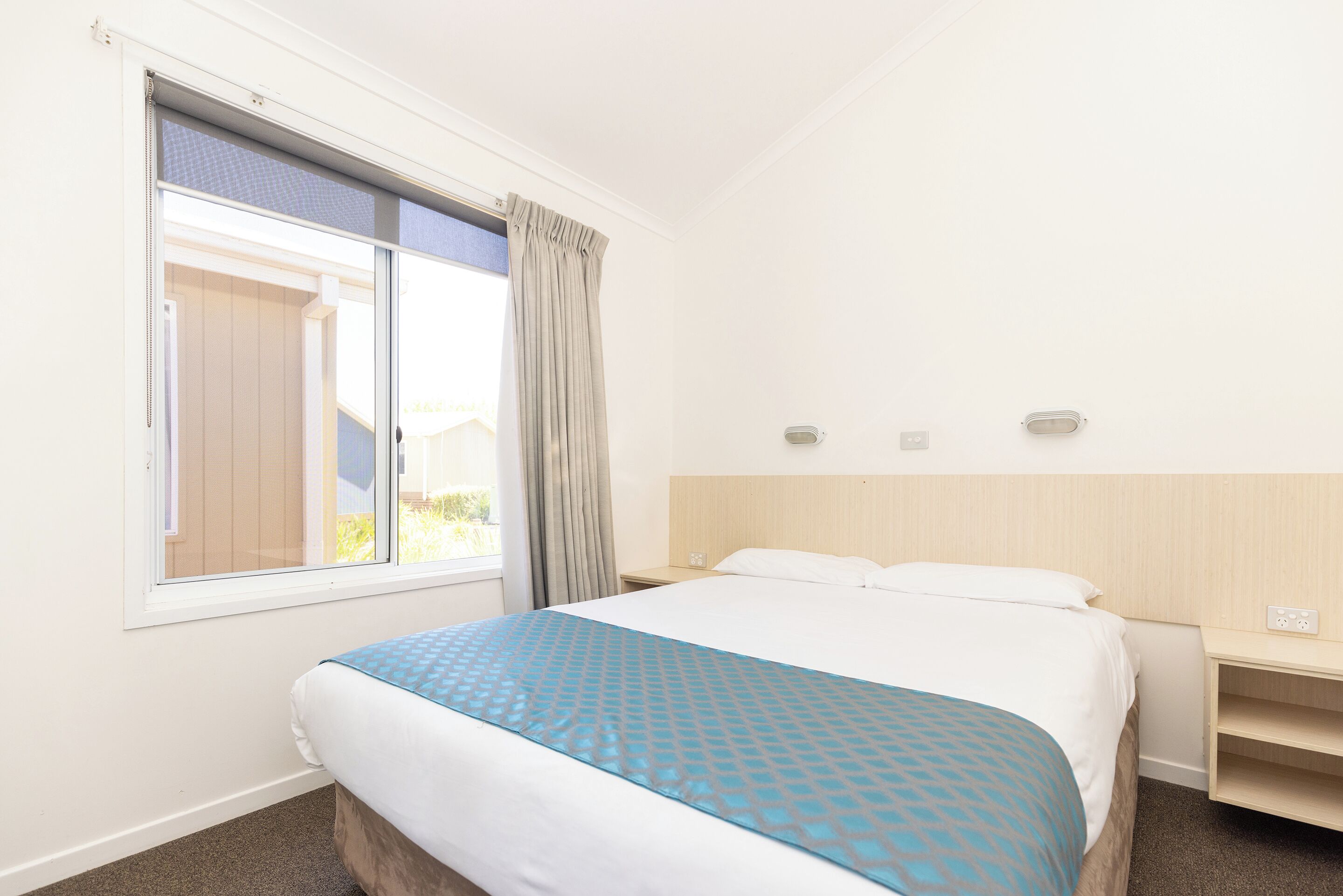 A hotel room with a bed, a window with curtains, a bedside table, and a wall-mounted light.