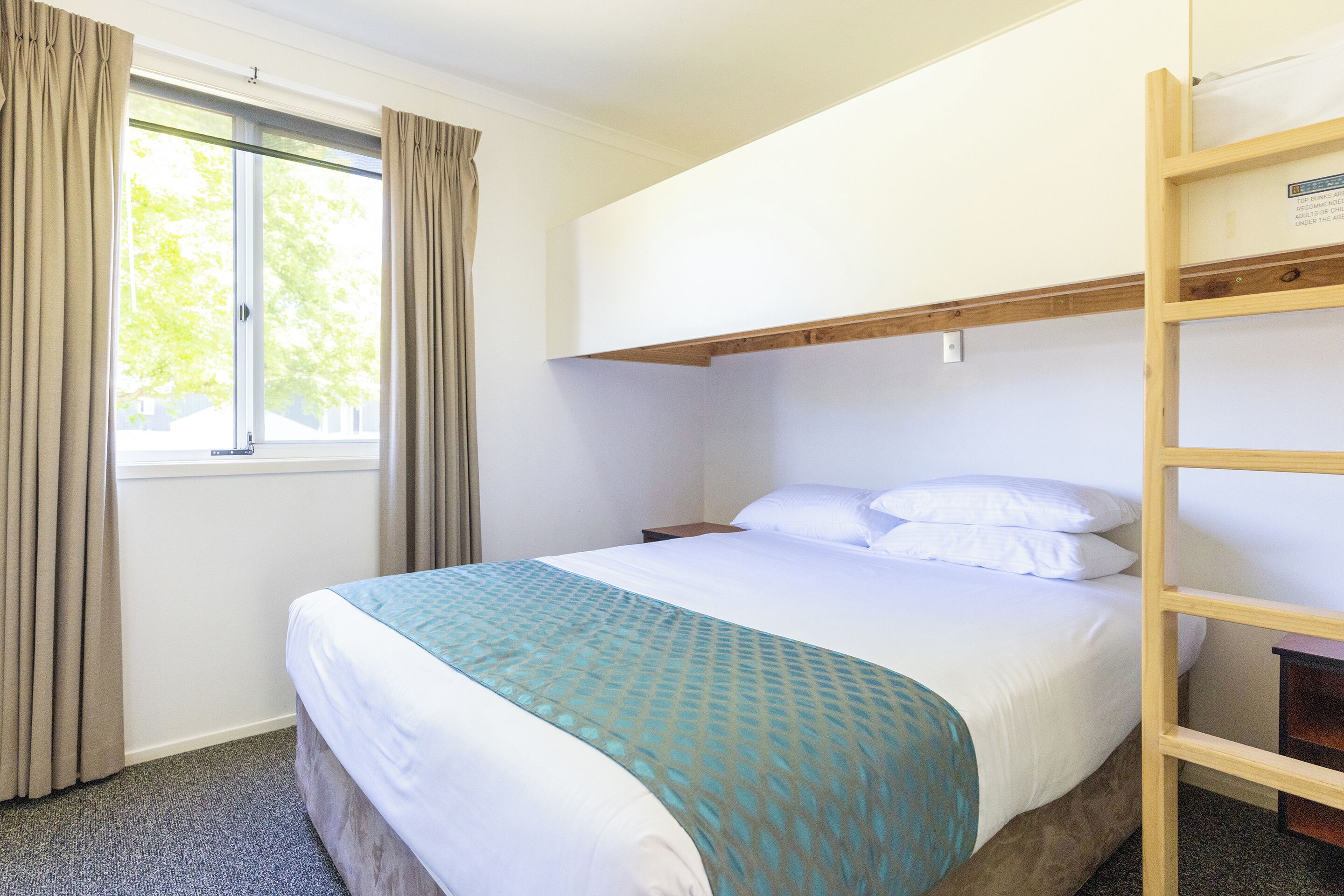 A hotel room with a bunk bed, a window with curtains, a bedside table, and a bed with a patterned blanket.