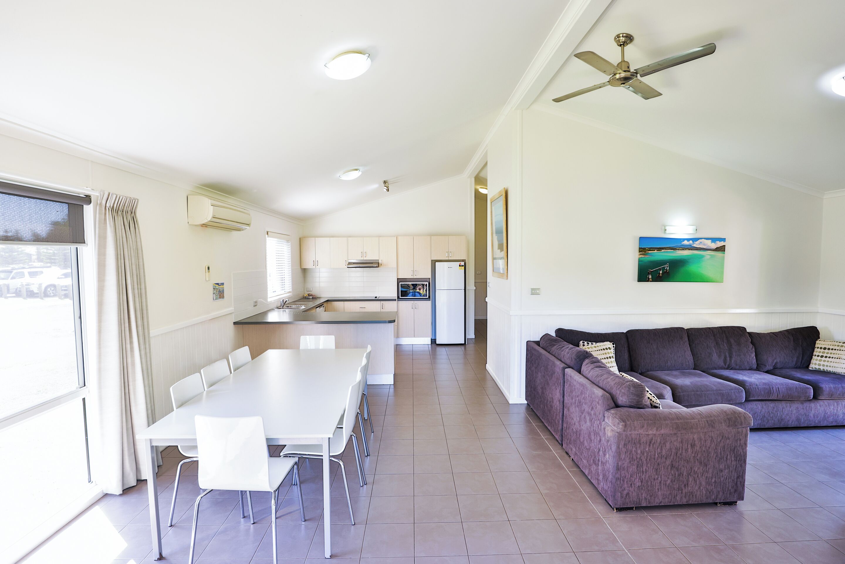 A modern kitchen with white cabinets, a dining table with chairs, and a purple sofa in a bright living area with a ceiling fan.