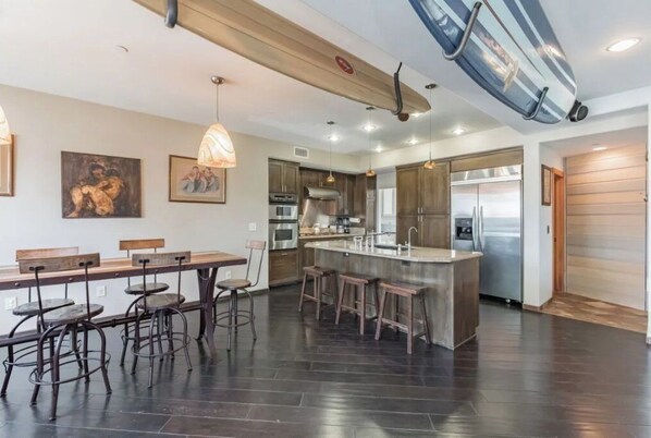 A modern kitchen with dark flooring, a central island with stools, stainless steel appliances, and a surfboard hanging from the ceiling.