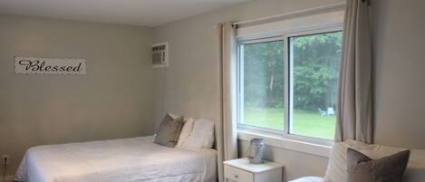 A bedroom with two beds, a bedside table with a lamp, a window with curtains, and a sign on the wall that says "Blessed".