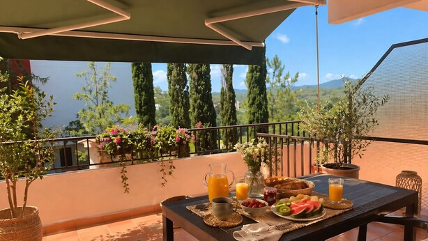 Un balcón con una mesa dispuesta para el desayuno, que incluye frutas, una jarra de jugo de naranja y una taza de café.