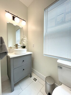 Une salle de bain avec un lavabo, un miroir et des toilettes blancs. Une serviette est accrochée au mur et une poubelle se trouve à côté des toilettes.