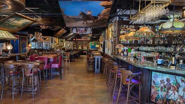 Eine Bar mit Holztischen und -stühlen, einem Wandgemälde und einer Auswahl an Flaschen und Gläsern auf Regalen.