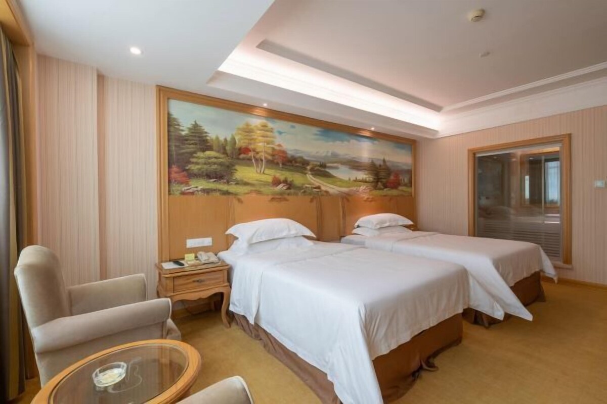 A hotel room with two beds, a desk, a chair, and a painting on the wall.