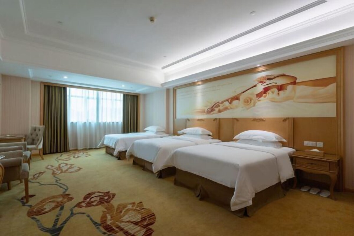 A hotel room with two beds, a sitting area with a sofa and chairs, a large painting on the wall, and a carpet with a floral design.
