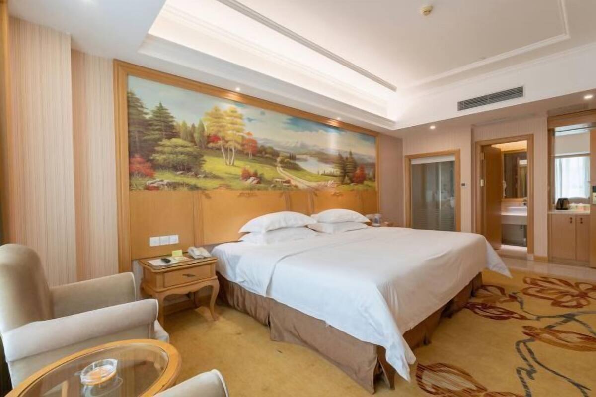 A hotel room with a large bed, a nightstand, a chair, a small table, and a painting on the wall.