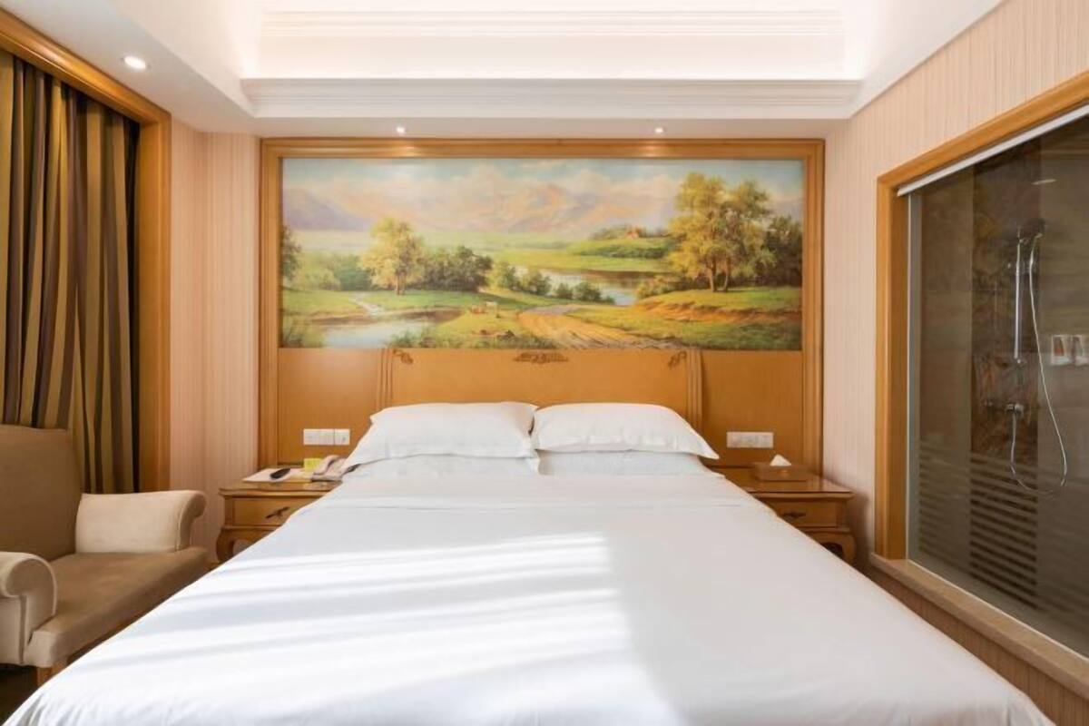 A hotel room with a large bed, a painting of a landscape, a chair, and a window with blinds.