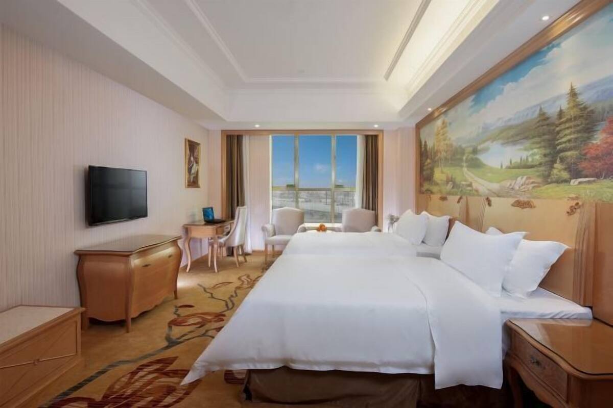 A hotel room with a large bed, a television, a desk, a chair, and a painting on the wall.