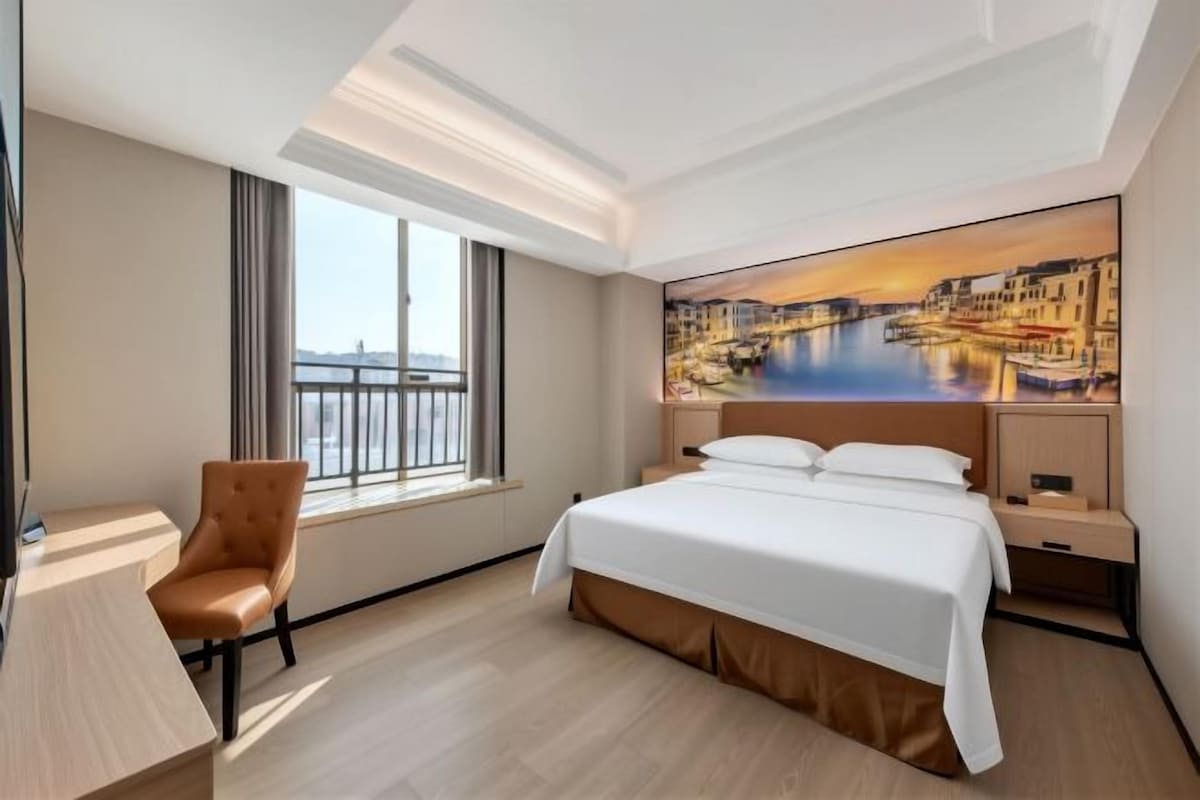 A hotel room with a bed, a desk, a chair, a window with a view, and a painting on the wall.