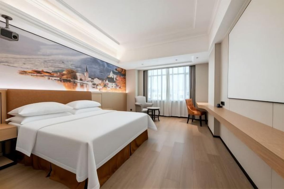 A hotel room with a large bed, a desk, a chair, and a cityscape painting on the wall.