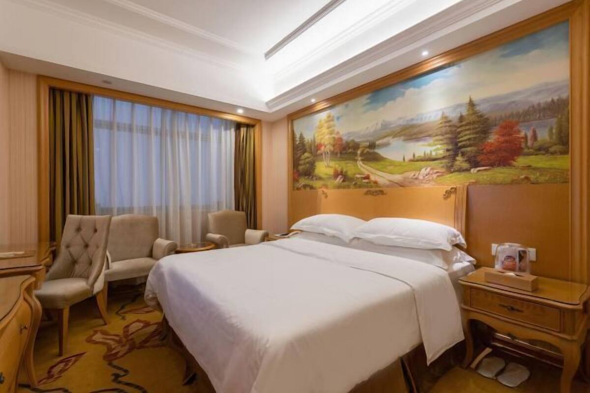 A hotel room with a large bed, a desk, a chair, and a painting of a landscape on the wall.