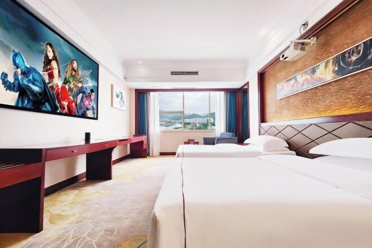 A hotel room with a large bed, a desk, a TV with a superhero movie, and a window with a city view.