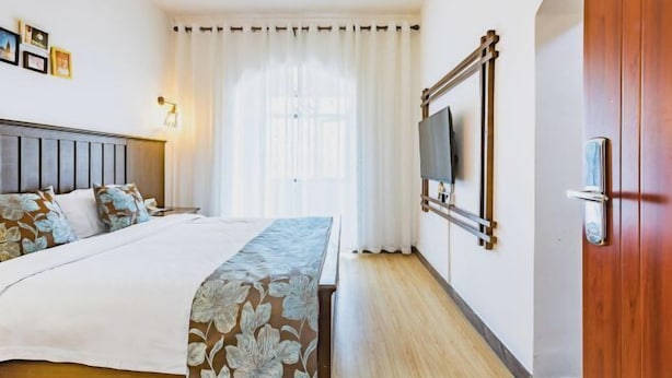 Hotellihuone, jossa on sänky, seinälle asennettu televisio, puuovi ja ikkuna, jossa on verhot.