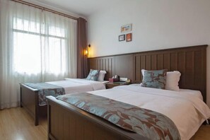 Habitación de hotel con dos camas, cabeceros de madera, una mesita de noche con lámpara y una ventana con cortinas translúcidas.