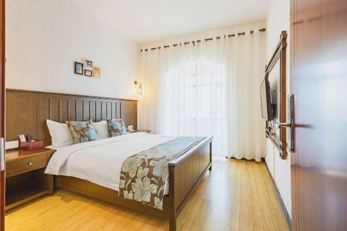 Un dormitorio con una cama de madera, una mesita de noche, una mesita junto a la cama, un televisor instalado en la pared y una ventana con cortinas.