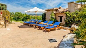 A patio with sun loungers and a white umbrella, surrounded by greenery and a stone wall.