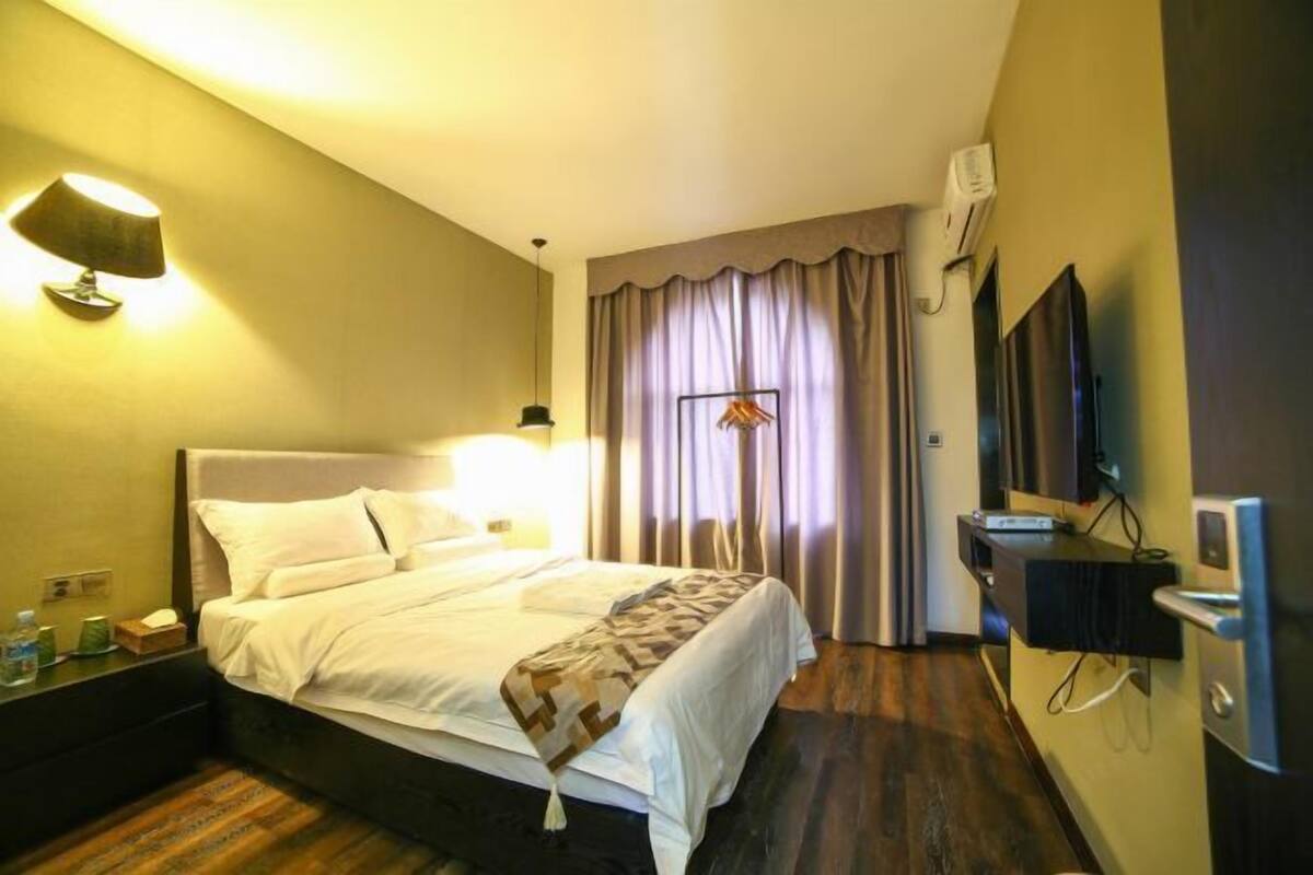 A hotel room with a bed, bedside table, lamp, curtains, TV, and a wall-mounted air conditioning unit.