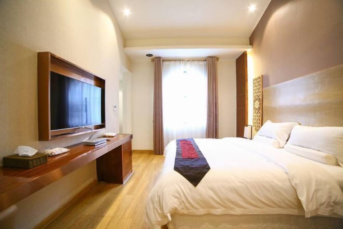 A hotel room with a bed, a TV on a wooden console, a window with curtains, and a wall-mounted artwork.