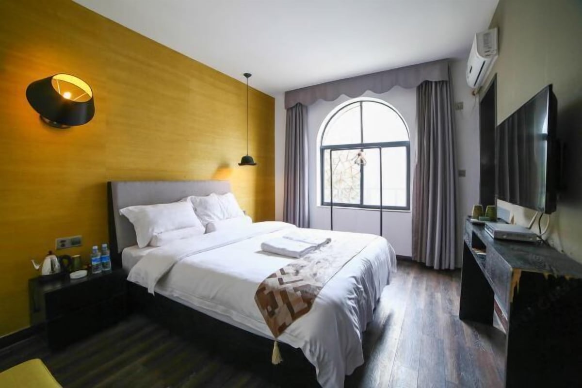 Ein Hotelzimmer mit Bett, Nachttisch, Lampe, Fernseher und einem großen Fenster mit Vorhängen.