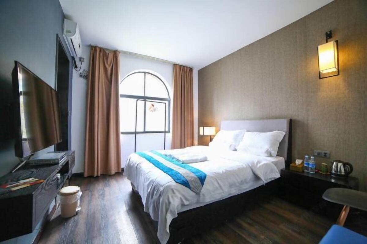 A hotel room with a bed, a desk, a television, a window with curtains, and a wall-mounted light fixture.