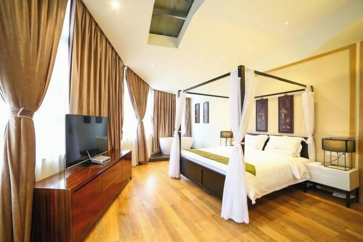 A bedroom with a four-poster bed, a TV on a wooden cabinet, and a large window with curtains.