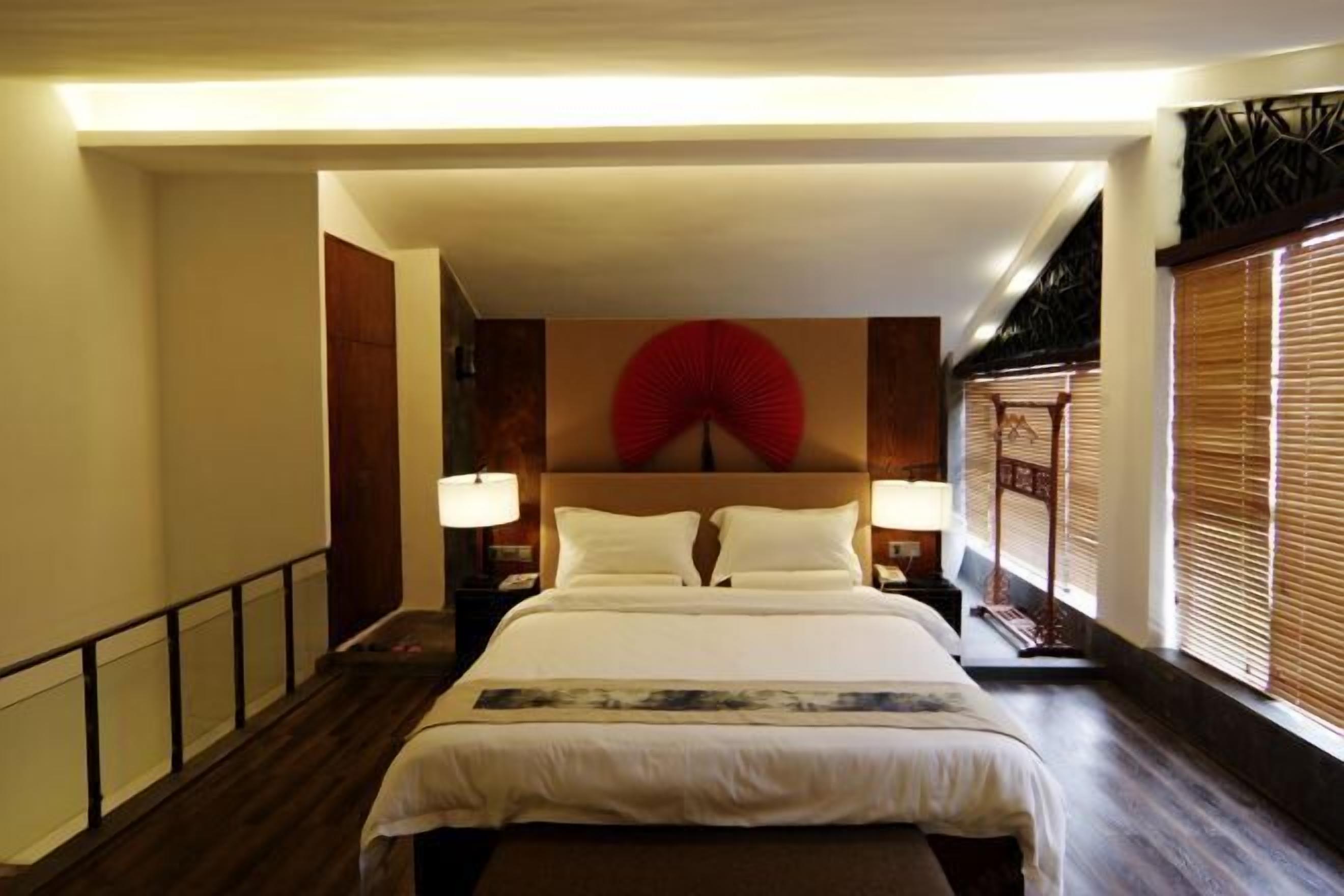 A hotel room with a large bed, bedside lamps, a wooden floor, and a window with blinds.