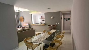 A modern living room with a sofa, a dining table with chairs, a kitchen area in the background, and a hallway leading to another room.