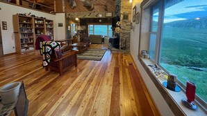Un salon spacieux doté de planchers en bois, d’une cheminée, d’une grande fenêtre offrant une vue sur un paysage verdoyant, ainsi que d’une bibliothèque remplie de livres et d’objets décoratifs.