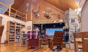 A cozy cabin interior with wooden floors, a stone fireplace, a bookshelf, and mounted deer heads.