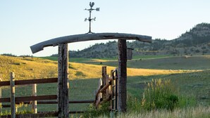 Une structure rustique en bois, surmontée d’une girouette, entourée d’une clôture en bois et bordée d’un champ d’herbe.