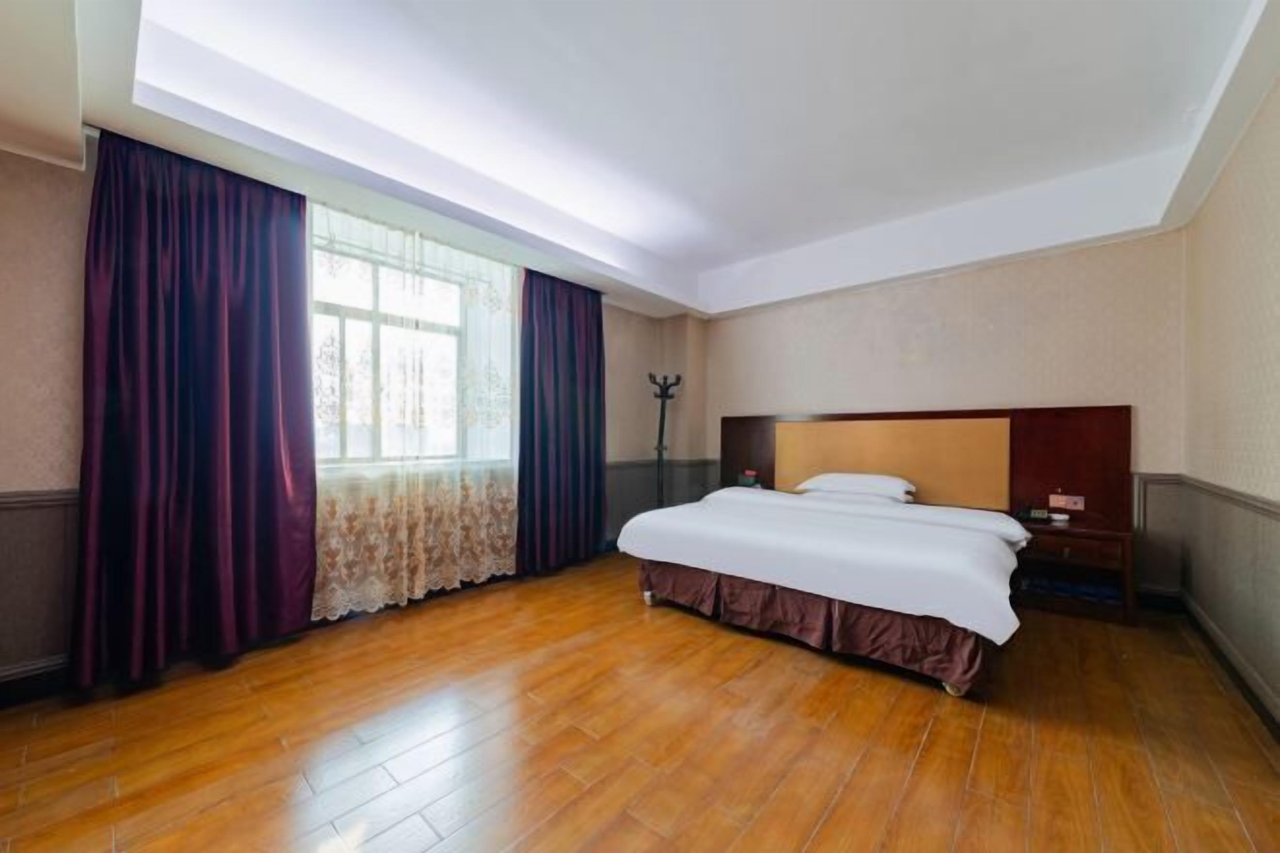 A hotel room with a large bed, wooden flooring, a window with curtains, and a nightstand.