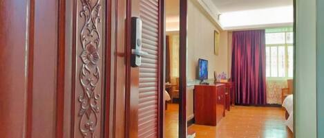 A hotel room with a wooden door, a desk with a computer, a bed, and a window with curtains.